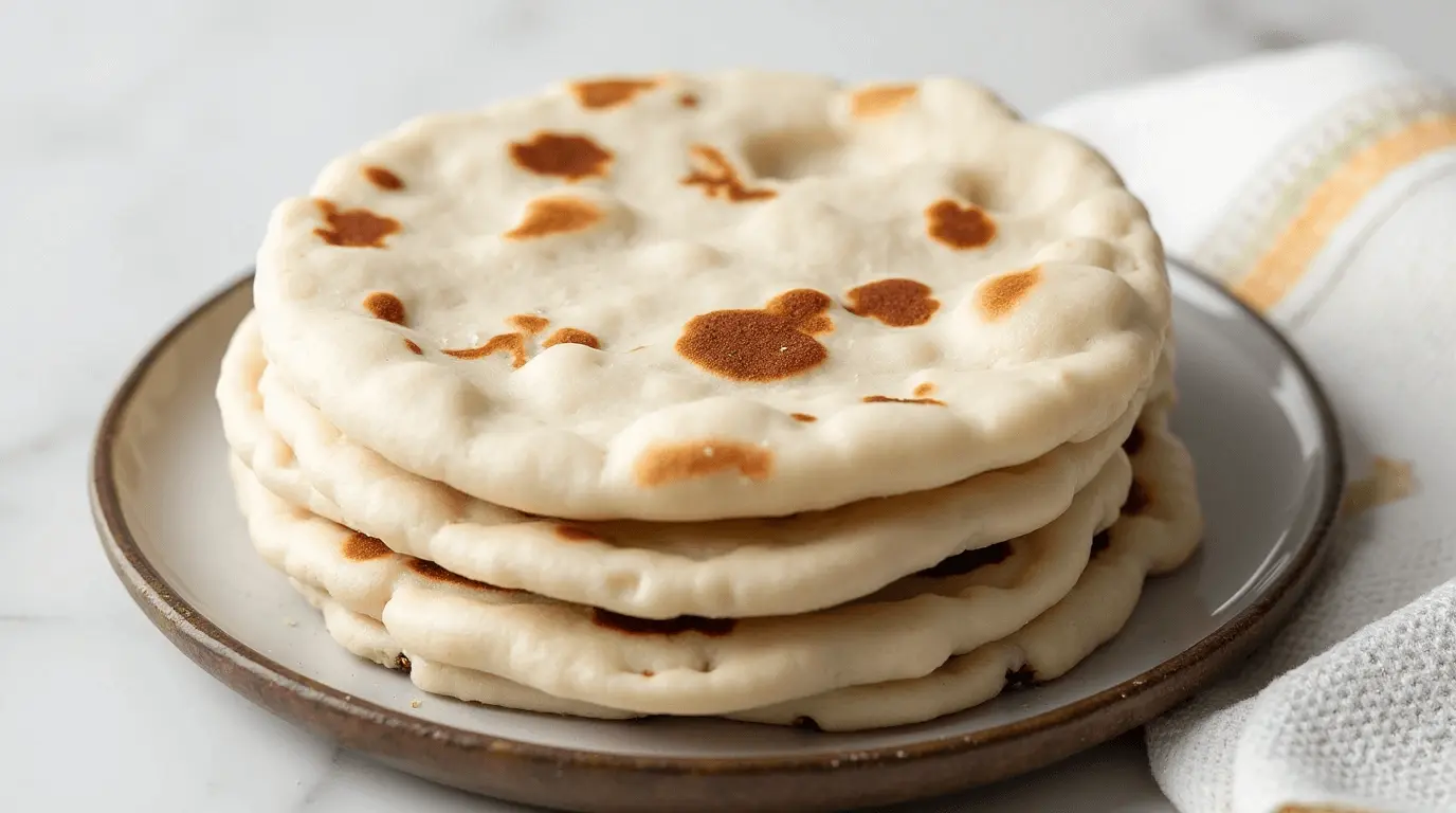 A stack of freshly cooked flatbreads with golden brown spots, served on a plate, accompanied by a folded kitchen towel in the background