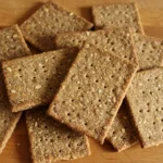 A close-up of homemade sourdough crackers with a golden-brown, crisp texture, topped with seeds, placed on a wooden cutting board.